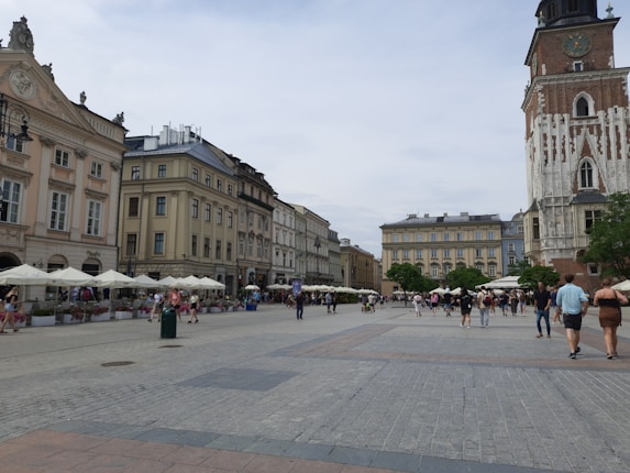 A bustling city square with historic architecture, featuring a range of buildings with ornate facades and a tall tower with a clock. The square is lined with outdoor cafes and people walking across the open cobblestone area, enjoying a leisurely afternoon.