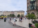 A bustling town square with people walking and gathering. Historic buildings with ornate architecture line one side, and a church with gothic elements is prominently featured. Yellow umbrellas cover outdoor seating areas, and greenery adds a touch of nature.