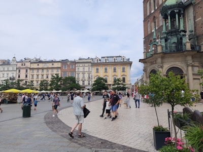 A bustling town square with people walking and gathering. Historic buildings with ornate architecture line one side, and a church with gothic elements is prominently featured. Yellow umbrellas cover outdoor seating areas, and greenery adds a touch of nature.