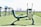An outdoor gym with several pieces of exercise equipment arranged on a green artificial turf. The image focuses on a leg press machine in the foreground with other fitness machines visible in the background. The area is surrounded by an open, empty landscape.