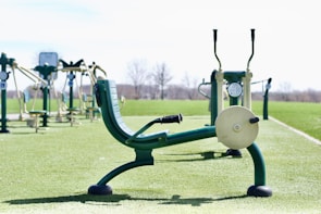 An outdoor gym with several pieces of exercise equipment arranged on a green artificial turf. The image focuses on a leg press machine in the foreground with other fitness machines visible in the background. The area is surrounded by an open, empty landscape.