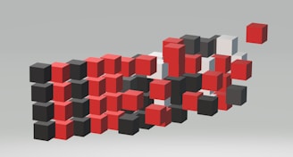 A collection of three-dimensional cubes in varying colors appears to be in motion or disarray. The cubes are primarily red, black, and white, and seem to be floating against a neutral, light gray background. The arrangement suggests a dynamic, abstract composition.