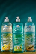 Three bottles of Earth Choice cleaning products are lined up on a wooden surface. The bottles have liquid inside and feature colorful labels with natural imagery like lemons and flowers. Each bottle is topped with a white cap, and the background is a solid teal color.