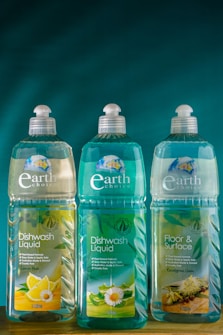 Three bottles of Earth Choice cleaning products are lined up on a wooden surface. The bottles have liquid inside and feature colorful labels with natural imagery like lemons and flowers. Each bottle is topped with a white cap, and the background is a solid teal color.