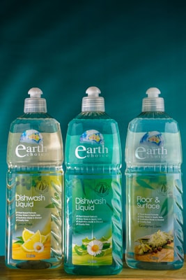 Three bottles of Earth Choice cleaning products are lined up on a wooden surface. The bottles have liquid inside and feature colorful labels with natural imagery like lemons and flowers. Each bottle is topped with a white cap, and the background is a solid teal color.