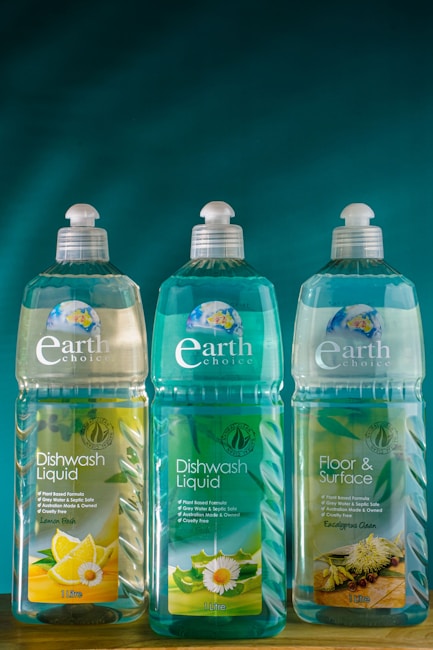 Three bottles of Earth Choice cleaning products are lined up on a wooden surface. The bottles have liquid inside and feature colorful labels with natural imagery like lemons and flowers. Each bottle is topped with a white cap, and the background is a solid teal color.