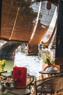Woven bamboo blinds hang partially down, casting shadows over a quaint outdoor seating area beside a tranquil river. Round glass-top tables are arranged with wicker chairs, one of which has a tea set on it. A red sign with Chinese characters stands prominently on a table. In the background, an arched stone bridge spans the river, and a few potted plants add greenery to the scene.