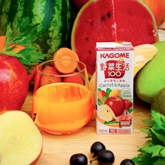 A variety of fresh fruits and vegetables surrounds a glass filled with orange juice. A carton labeled 'KAGOME Carrot & Apple' stands prominently. The assortment includes watermelon, apples, carrot slices, celery, black grapes, and other vibrant produce.