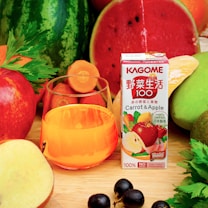 A variety of fresh fruits and vegetables surrounds a glass filled with orange juice. A carton labeled 'KAGOME Carrot & Apple' stands prominently. The assortment includes watermelon, apples, carrot slices, celery, black grapes, and other vibrant produce.