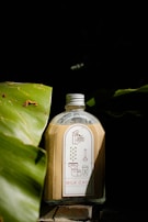 A bottle filled with a light brown liquid is placed between two large green leaves. The bottle has a white and red label with illustrations and the text 'Milk Cafe' and 'bean there'. The background is dark, creating a contrast with the bottle and leaves in the foreground.