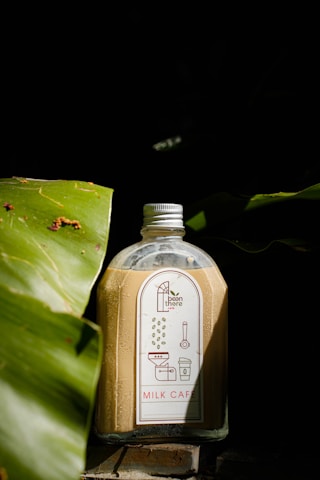 A bottle filled with a light brown liquid is placed between two large green leaves. The bottle has a white and red label with illustrations and the text 'Milk Cafe' and 'bean there'. The background is dark, creating a contrast with the bottle and leaves in the foreground.