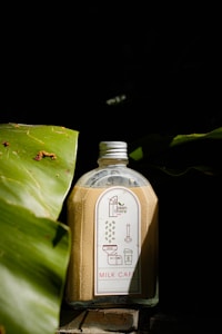 A bottle filled with a light brown liquid is placed between two large green leaves. The bottle has a white and red label with illustrations and the text 'Milk Cafe' and 'bean there'. The background is dark, creating a contrast with the bottle and leaves in the foreground.