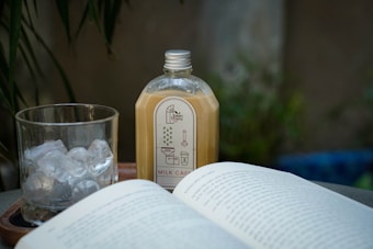 A bottle of milk tea labeled 'Milk Café' is placed on a wooden tray next to a glass filled with ice cubes. An open book with readable text is in the foreground. The setting appears to be outdoors, with green foliage in the background providing a blurred, natural backdrop.