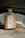 A bottle of milk coffee with a silver cap sits on a concrete surface. The label features illustrations of coffee beans, a grinder, and a coffee cup, with the text 'Bean There' and 'Milk Cafe'. An open book lies in the background, its pages illuminated by natural light.