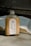 A bottle of milk coffee with a silver cap sits on a concrete surface. The label features illustrations of coffee beans, a grinder, and a coffee cup, with the text 'Bean There' and 'Milk Cafe'. An open book lies in the background, its pages illuminated by natural light.