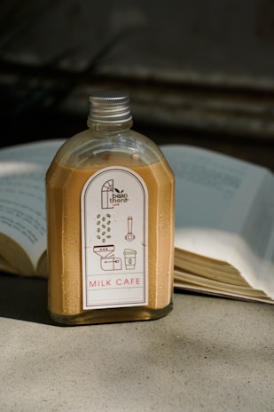 A bottle of milk coffee with a silver cap sits on a concrete surface. The label features illustrations of coffee beans, a grinder, and a coffee cup, with the text 'Bean There' and 'Milk Cafe'. An open book lies in the background, its pages illuminated by natural light.