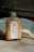 A bottle of milk coffee with a silver cap sits on a concrete surface. The label features illustrations of coffee beans, a grinder, and a coffee cup, with the text 'Bean There' and 'Milk Cafe'. An open book lies in the background, its pages illuminated by natural light.