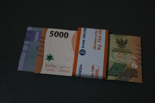 A stack of Indonesian banknotes featuring denominations of 5,000 and 10,000 Rupiah bound by a paper band indicating a bundled sum. The banknotes display various colors and design elements such as national emblems and geographic maps.