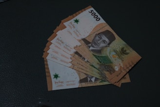 A stack of Indonesian 5000 Rupiah banknotes laid out on a dark surface. The notes feature a portrait along with intricate patterns and map details. The edges are fanned out to reveal multiple banknotes, showcasing their orange, green, and brown tones.