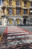 A yellow building with brown shutters and balconies positioned on a street corner. The ground level features a 'Ristorante' and 'Pizzeria' with closed yellow shutters. Multiple motorbikes are parked along the side of the road. Foreground includes a red and white pedestrian crossing.