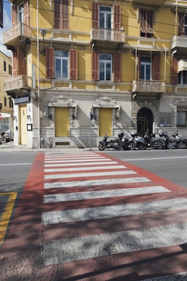 A yellow building with brown shutters and balconies positioned on a street corner. The ground level features a 'Ristorante' and 'Pizzeria' with closed yellow shutters. Multiple motorbikes are parked along the side of the road. Foreground includes a red and white pedestrian crossing.