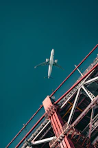An airplane is flying high in the sky above an architectural structure with red beams. The structure appears to be part of a bridge, with visible metal beams and cables. The composition includes a strong contrast between the white airplane and the vibrant red of the structure against a teal sky.