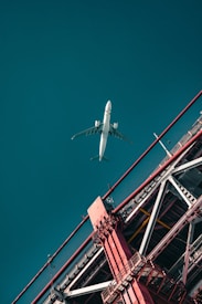 An airplane is flying high in the sky above an architectural structure with red beams. The structure appears to be part of a bridge, with visible metal beams and cables. The composition includes a strong contrast between the white airplane and the vibrant red of the structure against a teal sky.