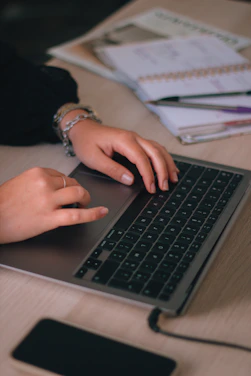 A close-up of hands typing on a laptop keyboard surrounded by books and notebooks.