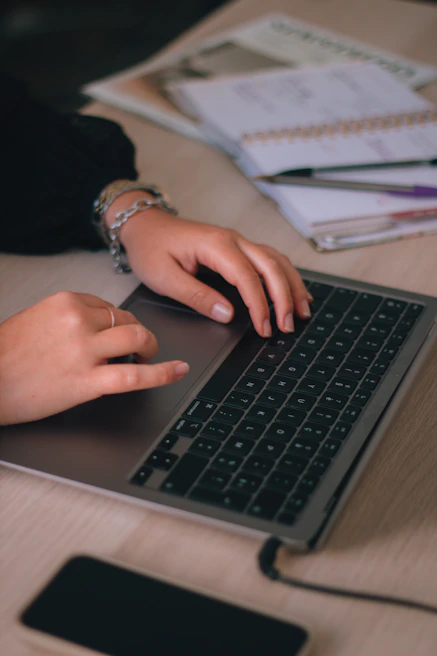 A close-up of hands typing on a keyboard with a blurred background of notes and a smartphone.