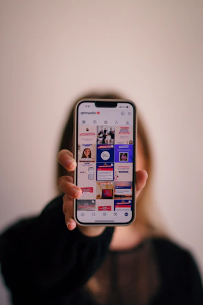 A person with long hair is holding a smartphone, displaying an Instagram profile with various posts. The background is softly blurred, emphasizing the phone.