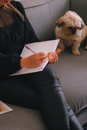 A person sitting on a couch is writing in a notebook with a pen. A small dog with fluffy fur is seated beside them, looking downwards. The setting appears cozy and intimate with soft, muted lighting.