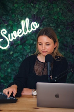 A woman sits at a wooden desk with a laptop in front of her. She has a thoughtful expression and is speaking into a microphone. The background features a lush, green leafy wall with the word 'Swello' illuminated in neon.