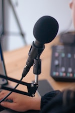 A close-up view of a microphone attached to an adjustable stand, with a hand typing on a laptop keyboard in the background. The setup suggests a recording or podcasting environment. A mixing console with colorful buttons can be partially seen, adding to the audio production context.