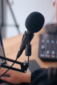 A close-up view of a microphone attached to an adjustable stand, with a hand typing on a laptop keyboard in the background. The setup suggests a recording or podcasting environment. A mixing console with colorful buttons can be partially seen, adding to the audio production context.