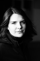 A black and white portrait of a person gazing directly into the camera. The image captures a serene and contemplative mood, with soft lighting creating gentle shadows on the face. The person is wearing a dark jacket and has medium-length hair that subtly moves with the light breeze.