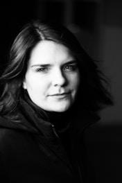 A black and white portrait of a person gazing directly into the camera. The image captures a serene and contemplative mood, with soft lighting creating gentle shadows on the face. The person is wearing a dark jacket and has medium-length hair that subtly moves with the light breeze.