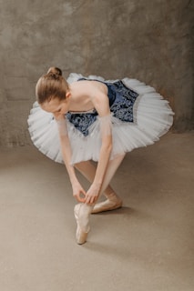 A ballerina is adjusting her pointe shoes. She is wearing a classical ballet tutu with a dark blue bodice and white tutu skirt. Her hair is neatly tied up in a bun, and she is standing on a neutral, textured surface with a gray background.