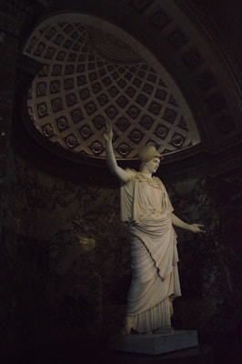 A marble statue of a classical figure, possibly a goddess, is positioned under an intricately patterned, domed ceiling. The statue is illuminated, contrasting against the darker background, creating a focus on its detailed drapery and serene expression. The surrounding area has marble walls that add an element of grandeur to the setting.