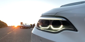 A close-up view of a white car's headlight, with the glowing rings prominently visible. In the background, there is a scenic road during a sunset, with a convertible sports car parked on the side and a few people standing near it.