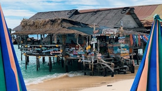 A rustic beachfront bar stands on stilts over clear turquoise water. The structure features thatched roofs and wooden planks adorned with a variety of colorful signs in different languages. The sandy beach and blue sky create a tropical atmosphere, while colorful umbrellas are visible in the foreground.