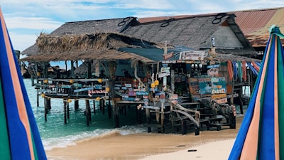 A rustic beachfront bar stands on stilts over clear turquoise water. The structure features thatched roofs and wooden planks adorned with a variety of colorful signs in different languages. The sandy beach and blue sky create a tropical atmosphere, while colorful umbrellas are visible in the foreground.