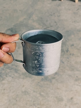 A hand holds a silver-colored metallic cup featuring intricate engraved patterns. The cup contains a liquid, likely water, and is being held against a neutral, slightly textured background.