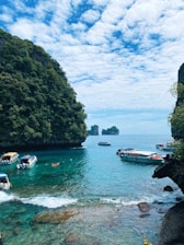 A scenic view of a tropical bay with clear blue water surrounded by lush green cliffs. Several boats are gently floating on the water, including small speedboats and a kayak. The sky is partly cloudy, adding texture to the bright, sunny day.