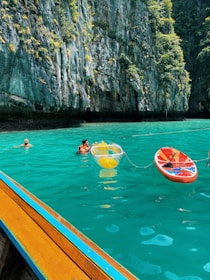 A serene body of turquoise water enclosed by tall, rugged cliffs with lush green vegetation. Three people are enjoying the water, two of them swimming while another appears to be holding onto an inflatable yellow and clear ball. There is also a red paddleboard nearby, and a part of a wooden boat with a blue stripe can be seen in the foreground.