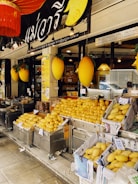 A fruit shop exterior is filled with heaps of ripe yellow mangoes displayed in baskets lined with newspapers. The store has a sign in a foreign script and large decorative mango figures are attached on either side. Bright lights illuminate the interior visible through the glass window.