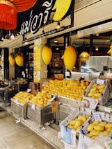 A fruit shop exterior is filled with heaps of ripe yellow mangoes displayed in baskets lined with newspapers. The store has a sign in a foreign script and large decorative mango figures are attached on either side. Bright lights illuminate the interior visible through the glass window.