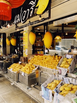 A fruit shop exterior is filled with heaps of ripe yellow mangoes displayed in baskets lined with newspapers. The store has a sign in a foreign script and large decorative mango figures are attached on either side. Bright lights illuminate the interior visible through the glass window.