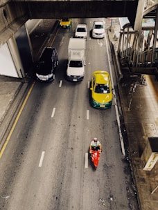 A top-down view of a multi-lane road with several vehicles including cars, a truck, and a motorcycle. The vehicles are mostly moving forward, with a yellow and green taxi and a red motorcycle standing out. The road is flanked by concrete sidewalks, and an overpass structure is visible in the upper part of the image.