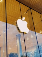 A brightly lit Apple logo mounted on the glass facade of a modern building with a wooden ceiling and metallic columns. Reflections and indoor plants are visible through the glass.