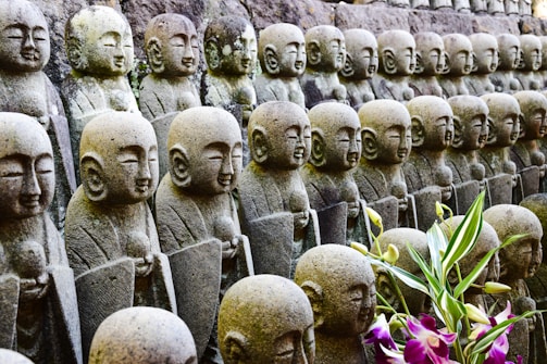 A row of small stone Buddhist statues with serene expressions are neatly aligned, likely representing Jizo, a protector figure. The statues are weathered and set against a rock background, with vibrant green leaves and purple and white flowers in the foreground.